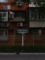 A urban residential building facade with bars on the windows and air conditioning units installed. A street sign in multiple languages is visible in the foreground, next to a fence and greenery along the sidewalk.