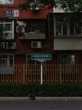 A urban residential building facade with bars on the windows and air conditioning units installed. A street sign in multiple languages is visible in the foreground, next to a fence and greenery along the sidewalk.