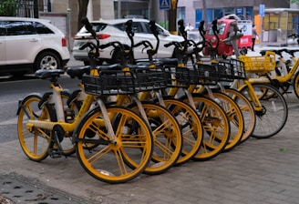 A row of yellow dockless rental bicycles is parked on a sidewalk. Each bike features a basket at the front and is aligned neatly. In the background, various vehicles including cars and a small red electric vehicle are visible on the adjacent street. The scene is urban and possibly taken in a city setting.