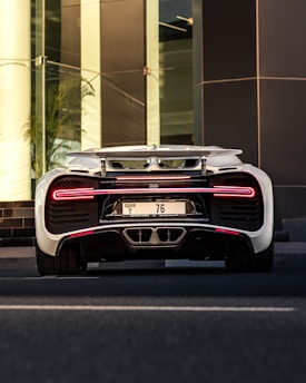 A high-performance sports car is captured from the rear end, showing its sleek and aerodynamic design. The vehicle has a white exterior with distinctive detailing and is stationed in front of a modern glass building.