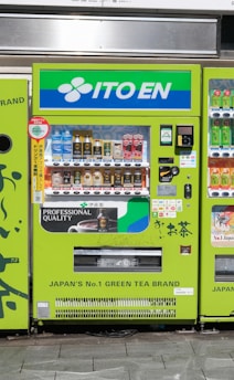 A bright green vending machine prominently displays the brand 'ITO EN' at the top. It offers a variety of beverages, including iced tea, coffee, and other drinks. The machine features a user-friendly interface with product images and prices. Branding and promotional details emphasize its association with a popular Japanese green tea brand.