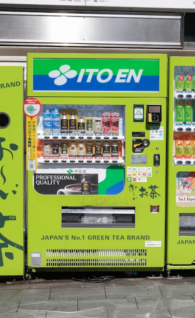 A bright green vending machine prominently displays the brand 'ITO EN' at the top. It offers a variety of beverages, including iced tea, coffee, and other drinks. The machine features a user-friendly interface with product images and prices. Branding and promotional details emphasize its association with a popular Japanese green tea brand.