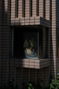 A mannequin styled like an ancient Egyptian pharaoh is placed inside a recessed window surrounded by a brick building facade. The pharaoh's iconic striped headdress is visible, containing hues of blue and gold. The facade comprises uniformly arranged bricks that create a geometric pattern. There are shadows cast across the bricks and the window, with some greenery at the base of the structure adding a touch of nature to the scene.