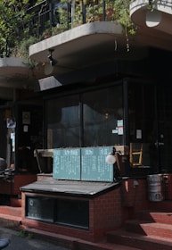 A quaint outdoor storefront features a menu board with Japanese writing, placed at the entrance of a cozy establishment. The entrance is made of dark glass panels and red brick, and there are metal kegs positioned nearby. Above the storefront, hanging planters with green foliage and ivy add a touch of nature.