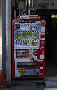 A bright red vending machine displaying various drinks including bottled water, teas, and sodas. The machine has advertisements for free Wi-Fi and gaming, along with several signs and labels in multiple languages. It is situated in an urban area, with a sidewalk partially visible beside it.