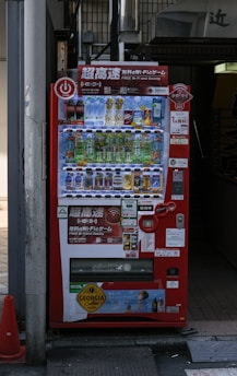 A bright red vending machine displaying various drinks including bottled water, teas, and sodas. The machine has advertisements for free Wi-Fi and gaming, along with several signs and labels in multiple languages. It is situated in an urban area, with a sidewalk partially visible beside it.