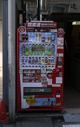 A bright red vending machine displaying various drinks including bottled water, teas, and sodas. The machine has advertisements for free Wi-Fi and gaming, along with several signs and labels in multiple languages. It is situated in an urban area, with a sidewalk partially visible beside it.