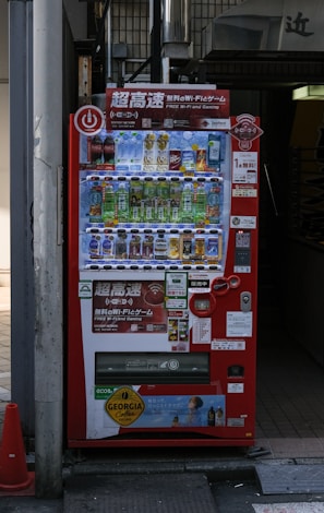 A bright red vending machine displaying various drinks including bottled water, teas, and sodas. The machine has advertisements for free Wi-Fi and gaming, along with several signs and labels in multiple languages. It is situated in an urban area, with a sidewalk partially visible beside it.