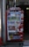 A bright red vending machine displaying various drinks including bottled water, teas, and sodas. The machine has advertisements for free Wi-Fi and gaming, along with several signs and labels in multiple languages. It is situated in an urban area, with a sidewalk partially visible beside it.