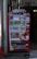A bright red vending machine displaying various drinks including bottled water, teas, and sodas. The machine has advertisements for free Wi-Fi and gaming, along with several signs and labels in multiple languages. It is situated in an urban area, with a sidewalk partially visible beside it.