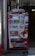 A bright red vending machine displaying various drinks including bottled water, teas, and sodas. The machine has advertisements for free Wi-Fi and gaming, along with several signs and labels in multiple languages. It is situated in an urban area, with a sidewalk partially visible beside it.