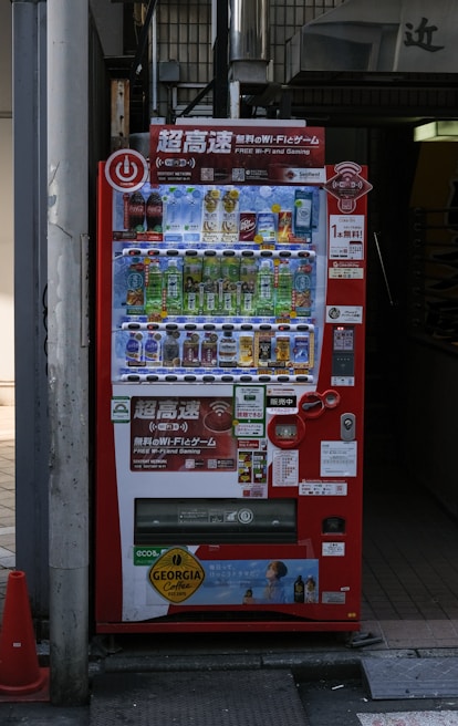 A bright red vending machine displaying various drinks including bottled water, teas, and sodas. The machine has advertisements for free Wi-Fi and gaming, along with several signs and labels in multiple languages. It is situated in an urban area, with a sidewalk partially visible beside it.