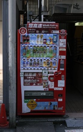 A bright red vending machine displaying various drinks including bottled water, teas, and sodas. The machine has advertisements for free Wi-Fi and gaming, along with several signs and labels in multiple languages. It is situated in an urban area, with a sidewalk partially visible beside it.