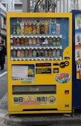A bright yellow vending machine filled with a variety of beverages including canned sodas, juices, and teas. The machine is placed on an urban sidewalk with signage and colorful labels in Japanese. A notable feature is its cheerful branding with cartoon characters and logos. The immediate background includes elements of a city street, some advertisements, and architectural details.
