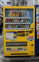A bright yellow vending machine filled with a variety of beverages including canned sodas, juices, and teas. The machine is placed on an urban sidewalk with signage and colorful labels in Japanese. A notable feature is its cheerful branding with cartoon characters and logos. The immediate background includes elements of a city street, some advertisements, and architectural details.
