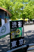 A signpost with Japanese characters and a cartoon image of a bird wearing a headband. It's located in an outdoor area with lush green trees and yellow tents in the background. A part of a building with blue and brown colors is visible on the left side.