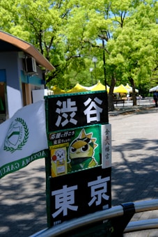 A signpost with Japanese characters and a cartoon image of a bird wearing a headband. It's located in an outdoor area with lush green trees and yellow tents in the background. A part of a building with blue and brown colors is visible on the left side.