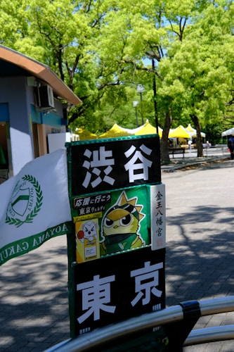 A signpost with Japanese characters and a cartoon image of a bird wearing a headband. It's located in an outdoor area with lush green trees and yellow tents in the background. A part of a building with blue and brown colors is visible on the left side.