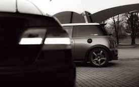 Two cars are parked under a tent-like structure. The foreground car is slightly blurred, while the background car, a compact model, is in focus. The scene is set in an outdoor area with some trees visible in the distance. The image has a monochrome filter, giving it a classic and nostalgic feel.