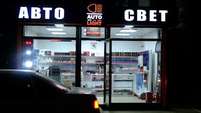 A storefront illuminated at night displays signage in Cyrillic, suggesting an automotive-related shop. The interior is brightly lit, showcasing shelves stocked with various automotive products and accessories. A car is parked in front of the glass window.