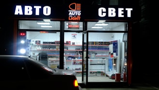 A storefront illuminated at night displays signage in Cyrillic, suggesting an automotive-related shop. The interior is brightly lit, showcasing shelves stocked with various automotive products and accessories. A car is parked in front of the glass window.