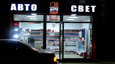 A storefront illuminated at night displays signage in Cyrillic, suggesting an automotive-related shop. The interior is brightly lit, showcasing shelves stocked with various automotive products and accessories. A car is parked in front of the glass window.