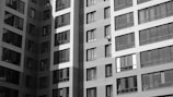A monochromatic image featuring the facade of a modern high-rise building with numerous windows. The architectural design includes vertical and horizontal line patterns, enhancing the visual texture. Some windows have reflections, while others appear dark, suggesting that the photograph was taken in natural light with shadows cast across the surface.