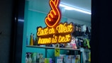 A neon sign with a peace hand gesture above glowing text that reads 'East or West home is best'. Behind the sign, a shop window display contains various colorful figurines and some retail products like bottles and packaged goods. The shop interior has teal-colored walls and shelves with hanging merchandise.