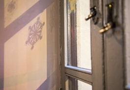 A close-up view of a window with frosted glass. The glass features a decorative snowflake pattern, and there are shadows and reflections visible. The window frame is metallic, and there is a latch partly visible.