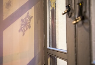 A close-up view of a window with frosted glass. The glass features a decorative snowflake pattern, and there are shadows and reflections visible. The window frame is metallic, and there is a latch partly visible.