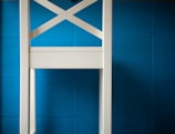 A white wooden chair with a cross-back design is positioned against a bright blue tiled wall. The chair's structure is simple and modern, with clean lines and a smooth finish. The background consists of evenly distributed blue tiles, creating a vibrant contrast with the chair.