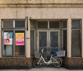 A vintage storefront features a closed double door and multiple frosted glass windows framed by beige tiled walls with dark brown accents at the base. A cream-colored bicycle fitted with two baskets is parked in front of the door. There are two posters on the left glass window displaying text and photos.