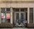 A vintage storefront features a closed double door and multiple frosted glass windows framed by beige tiled walls with dark brown accents at the base. A cream-colored bicycle fitted with two baskets is parked in front of the door. There are two posters on the left glass window displaying text and photos.