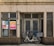 A vintage storefront features a closed double door and multiple frosted glass windows framed by beige tiled walls with dark brown accents at the base. A cream-colored bicycle fitted with two baskets is parked in front of the door. There are two posters on the left glass window displaying text and photos.