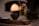 A clear glass filled with olive oil stands on a rustic wooden surface. Inside the glass, a brush is soaking in the oil. A small dark ceramic bowl is visible beside the glass, all set against a blurred background with warm lighting that suggests an intimate, cozy atmosphere.