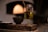 A clear glass filled with olive oil stands on a rustic wooden surface. Inside the glass, a brush is soaking in the oil. A small dark ceramic bowl is visible beside the glass, all set against a blurred background with warm lighting that suggests an intimate, cozy atmosphere.