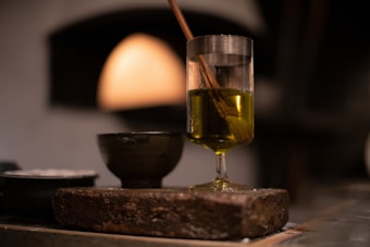 A clear glass filled with olive oil stands on a rustic wooden surface. Inside the glass, a brush is soaking in the oil. A small dark ceramic bowl is visible beside the glass, all set against a blurred background with warm lighting that suggests an intimate, cozy atmosphere.