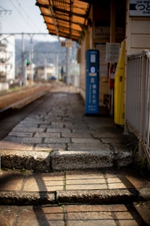 A deserted train platform with stone paving and a small ticket machine in a rural area. The platform has a metal roof with a yellowish tint, casting shadows on the ground. In the background, there are railway tracks stretching into the distance, flanked by power lines and blurred greenery.