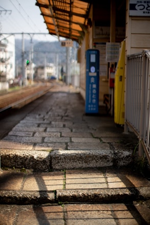 A deserted train platform with stone paving and a small ticket machine in a rural area. The platform has a metal roof with a yellowish tint, casting shadows on the ground. In the background, there are railway tracks stretching into the distance, flanked by power lines and blurred greenery.