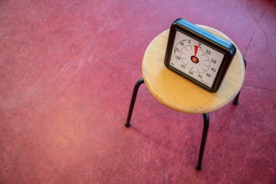 A small, wooden stool with black metal legs is placed on a red floor. On the stool, there is a square-shaped black timer displaying time with a white face and black numeral markings. The timer is set to around 55 minutes.