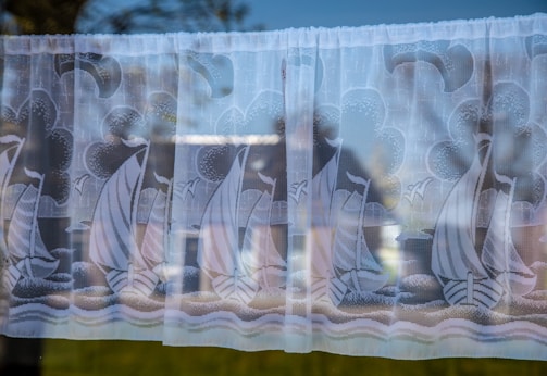 A close-up view of a lace curtain with an intricate design featuring sailboats and nautical themes. The curtain is hanging in front of a window, with a faint reflection of outdoor scenery visible through the glass.