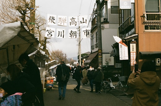 A bustling street market scene with people walking along a road lined with stalls and shops. Banners with Japanese writing are suspended between buildings. Several pedestrians are visible, some browsing the stalls, while others are taking photos or walking. The area has an old-town charm with traditional and modern elements.