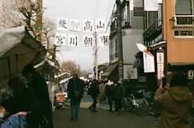 A bustling street market scene with people walking along a road lined with stalls and shops. Banners with Japanese writing are suspended between buildings. Several pedestrians are visible, some browsing the stalls, while others are taking photos or walking. The area has an old-town charm with traditional and modern elements.