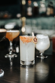 A beautifully arranged collection of cocktails set on a dark wooden table in a sophisticated bar setting. The focus is on a tall glass filled with a light-colored drink, garnished with a slice of dried citrus and a rim coated in chili powder or similar seasoning. Surrounding it are other cocktails in various shapes of glasses, each with a different color and garnish, creating an inviting and upscale atmosphere.