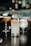 A beautifully arranged collection of cocktails set on a dark wooden table in a sophisticated bar setting. The focus is on a tall glass filled with a light-colored drink, garnished with a slice of dried citrus and a rim coated in chili powder or similar seasoning. Surrounding it are other cocktails in various shapes of glasses, each with a different color and garnish, creating an inviting and upscale atmosphere.