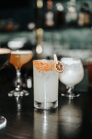 A beautifully arranged collection of cocktails set on a dark wooden table in a sophisticated bar setting. The focus is on a tall glass filled with a light-colored drink, garnished with a slice of dried citrus and a rim coated in chili powder or similar seasoning. Surrounding it are other cocktails in various shapes of glasses, each with a different color and garnish, creating an inviting and upscale atmosphere.