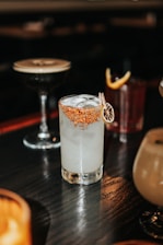 A variety of cocktails are placed on a dark wooden table. The central cocktail in a clear glass is filled with ice and has a red, spicy rim garnished with a slice of dried citrus. In the background, there are other cocktails, including a dark drink in a coupe glass and another in a tumbler with a lemon twist.