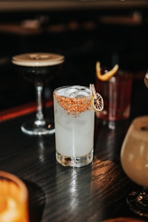 A variety of cocktails are placed on a dark wooden table. The central cocktail in a clear glass is filled with ice and has a red, spicy rim garnished with a slice of dried citrus. In the background, there are other cocktails, including a dark drink in a coupe glass and another in a tumbler with a lemon twist.