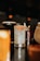 A glass filled with a light-colored cocktail, garnished with chili flakes on the rim and a dried citrus wheel. The background is dimly lit with bokeh lights, and other blurred glasses can be seen.