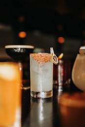 A glass filled with a light-colored cocktail, garnished with chili flakes on the rim and a dried citrus wheel. The background is dimly lit with bokeh lights, and other blurred glasses can be seen.